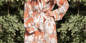 Woman standing hand on hip in long orange robe(?) in front of trees.