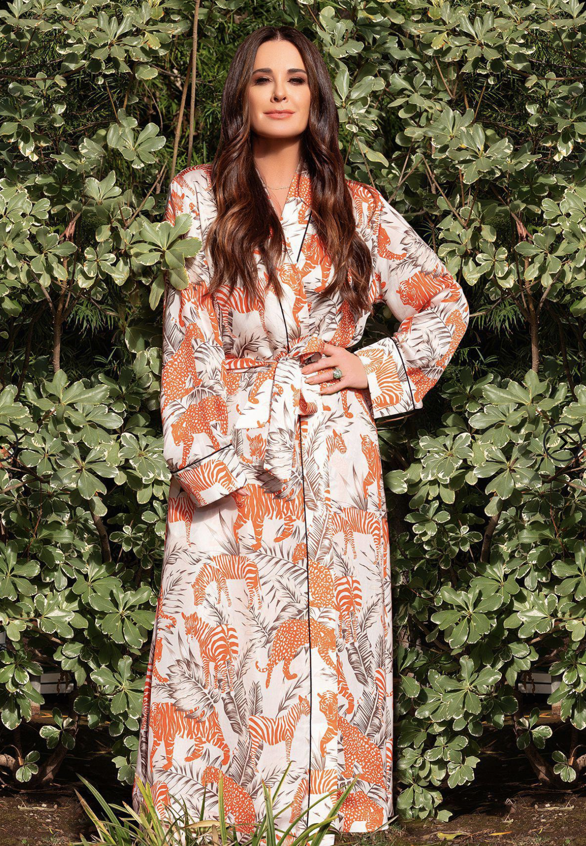 Woman standing hand on hip in long orange robe(?) in front of trees.